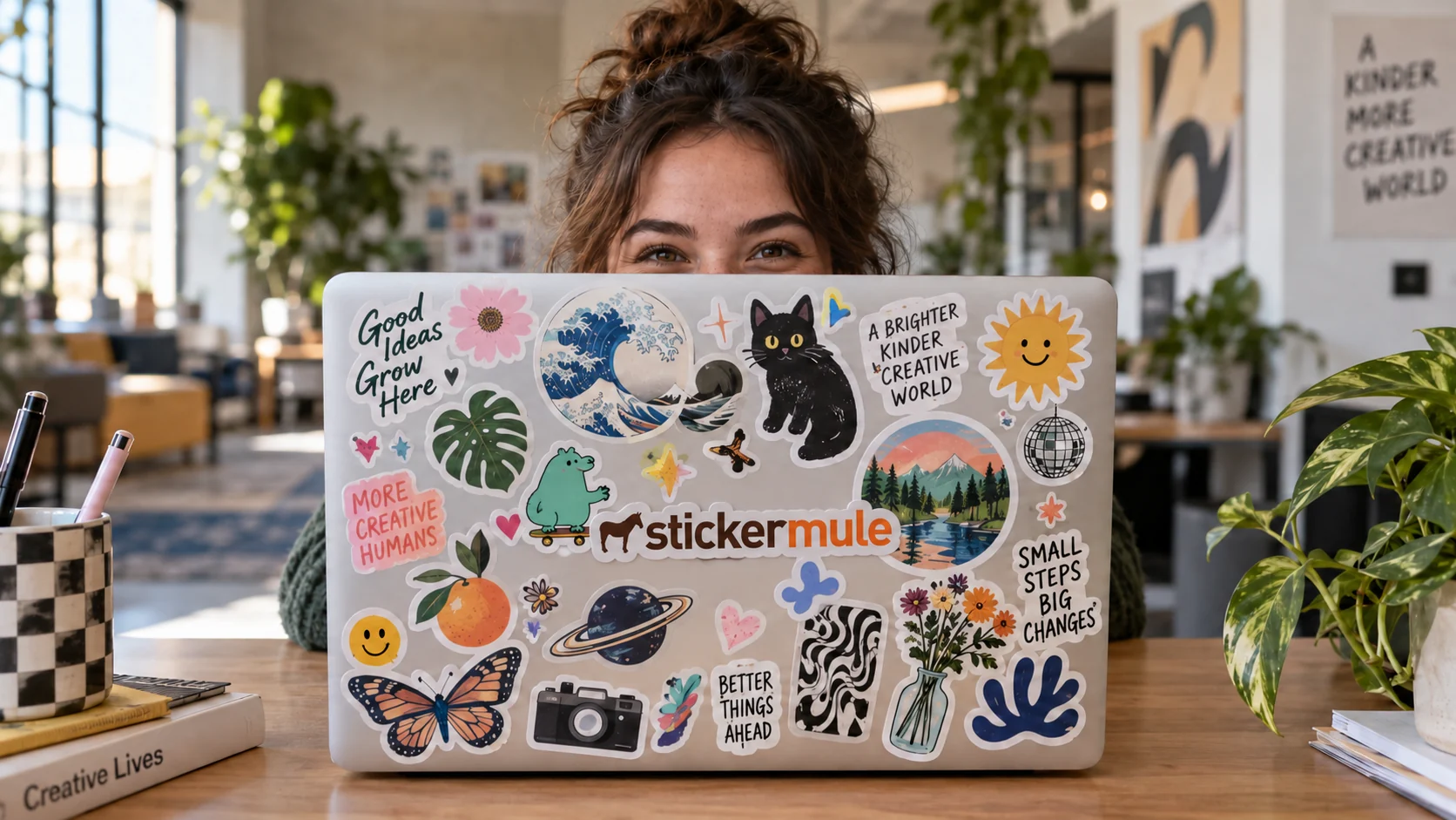 A young adult peers over a MacBook covered in colorful stickers, including a Sticker Mule logo, in a creative office.